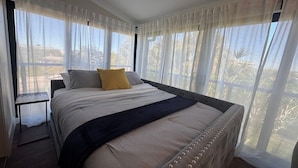 A bedroom with a large bed, sheer curtains, and a view of palm trees.