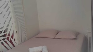 Ein Einzelbett mit weißer, strukturierter Bettdecke und zwei Kissen, ein aufgerolltes weißes Handtuch und eine weiße Trennwand mit geometrischem Muster.