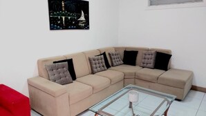 Un sofá seccional color beige con cojines negros y grises, una mesa de centro de vidrio con una vela blanca, y un cuadro con fuegos artificiales en la pared.