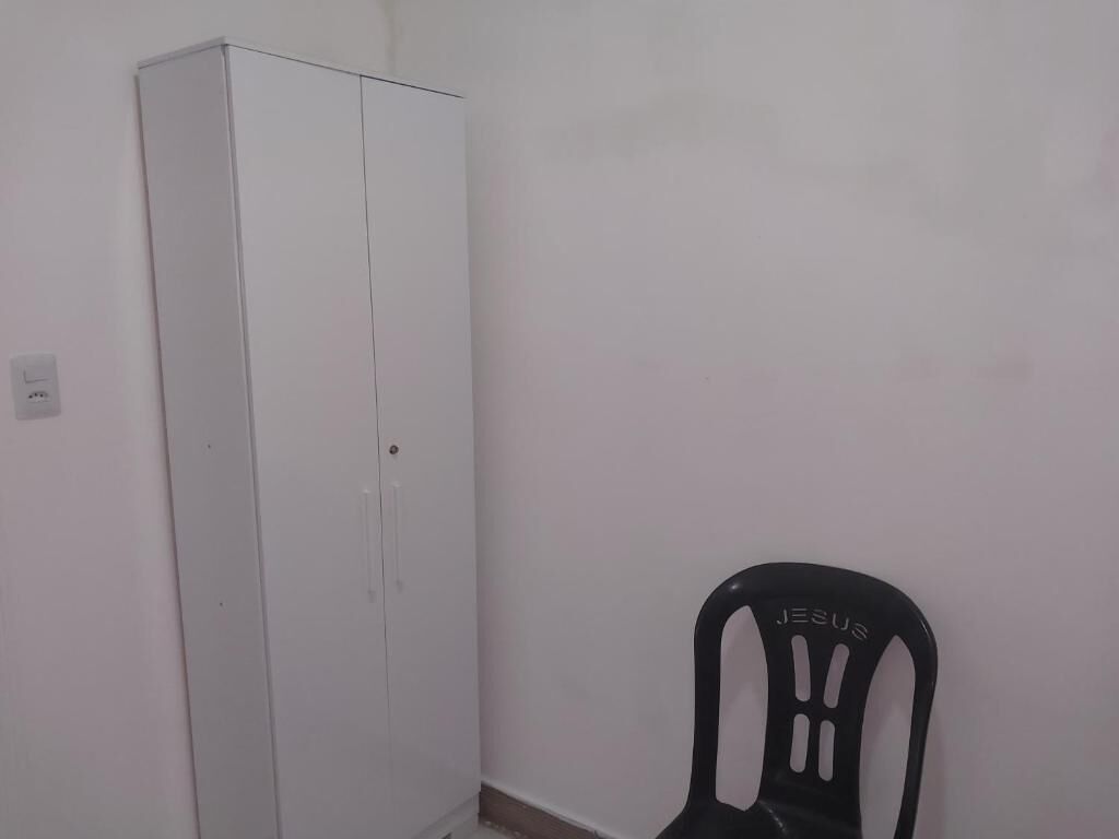 Quarto com um guarda-roupa branco e uma cadeira de plástico preta com a inscrição 'JESUS'.
