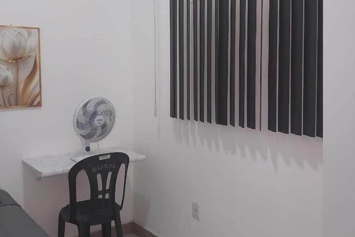 Una habitación pequeña con una cama, un escritorio con silla, un ventilador de pie y una ventana con persianas verticales.