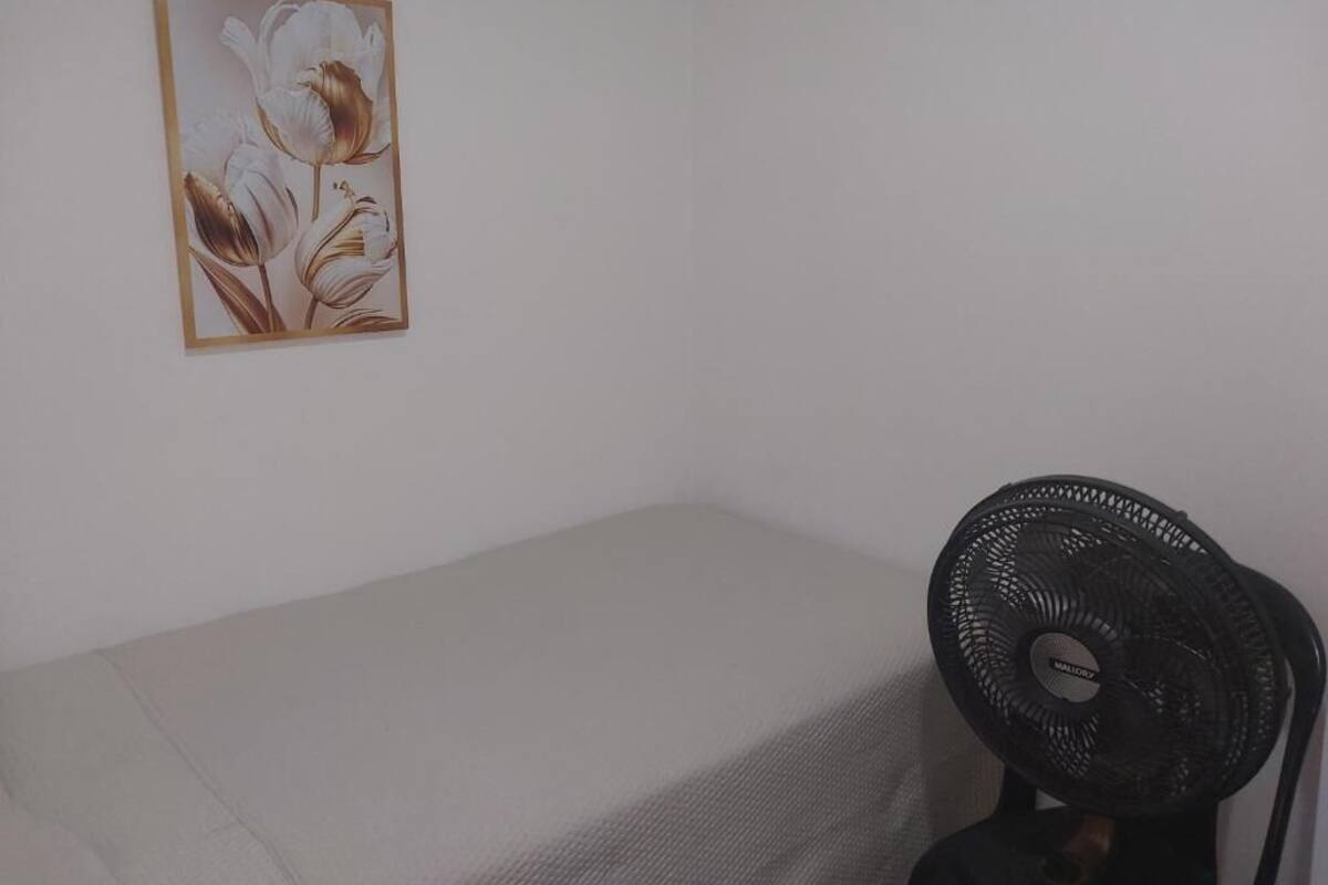 Una habitación con una cama, un cuadro de flores colgado en la pared y un ventilador de pie.