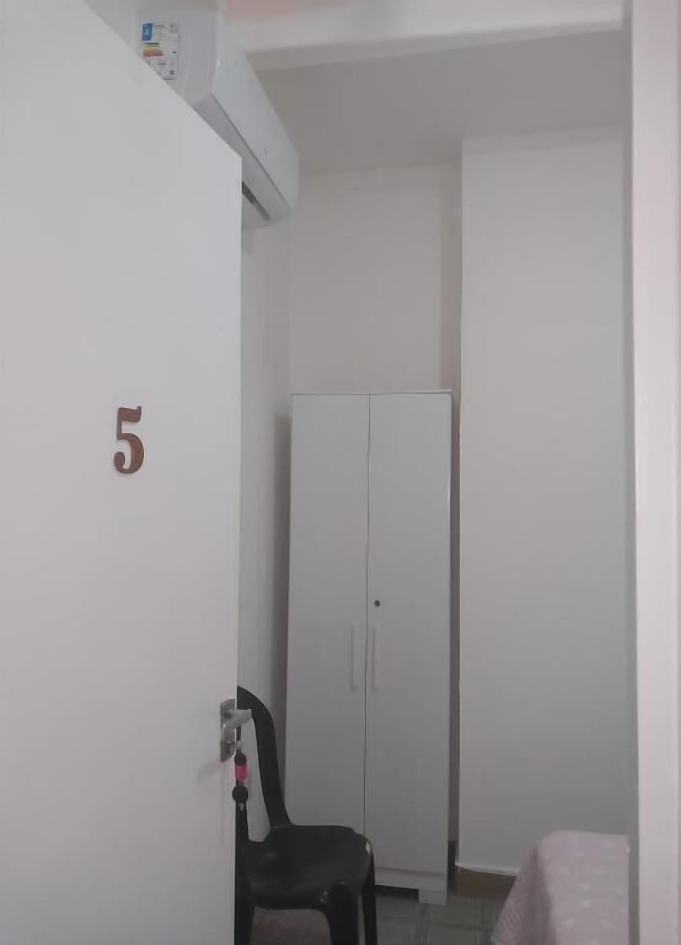 Una habitación con una puerta blanca numerada con el número 5, un armario blanco y una silla negra.