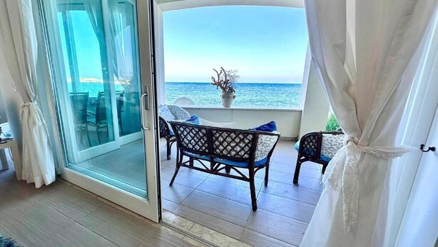 A room with a sliding glass door leading to a balcony with a view of the sea. There are two wicker chairs and a small table with a vase of flowers. - Casa Alba Serena by the sea (Marzamemi)