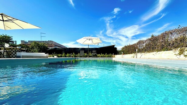 Piscina azul cristalina con sombrillas y sillas de descanso, bordeada por una playa de arena y árboles.