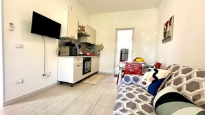 Una sala moderna con sofá, televisor de pantalla plana montado en la pared y una cocina con armarios y microondas.