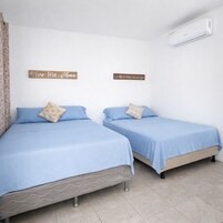 Dos camas individuales con ropa de cama azul y almohadas color beige, un equipo de aire acondicionado blanco en la pared, y dos letreros de madera con texto en español.