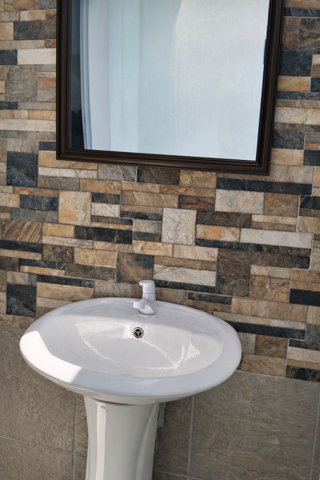 A white pedestal sink with a modern faucet, set against a wall with a mosaic of stone tiles.