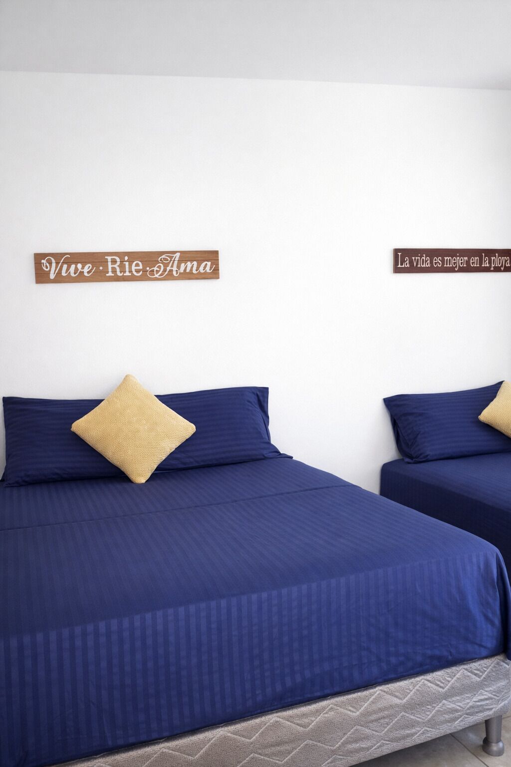 Dos camas con ropa de cama azul y un cojín beige, una pared blanca y dos letreros de madera con texto.