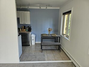 A compact kitchen with white cabinets, a coffee maker, and a trash bin. A small dining table with plates and a vase of dried flowers is placed near a window. - Awesome apartment in Seattle at walking distance from SCC - free parking. (Seattle)
