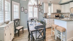 A kitchen with white cabinets, a dining table with chairs, a chandelier, and a large window with blinds. - Spectacular Southend 3BR Condo • Sleeps 7 • Rooftop Deck & Parking (Ocean City)