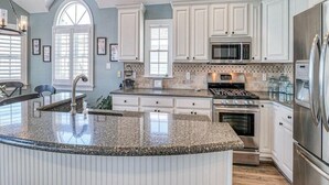 A kitchen with white cabinets, a granite countertop, stainless steel appliances, and a large window. - Spectacular Southend 3BR Condo • Sleeps 7 • Rooftop Deck & Parking (Ocean City)