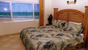 Quarto com cama, janela com vista para o oceano, cabeceira de madeira e mesa de cabeceira pequena com abajur.