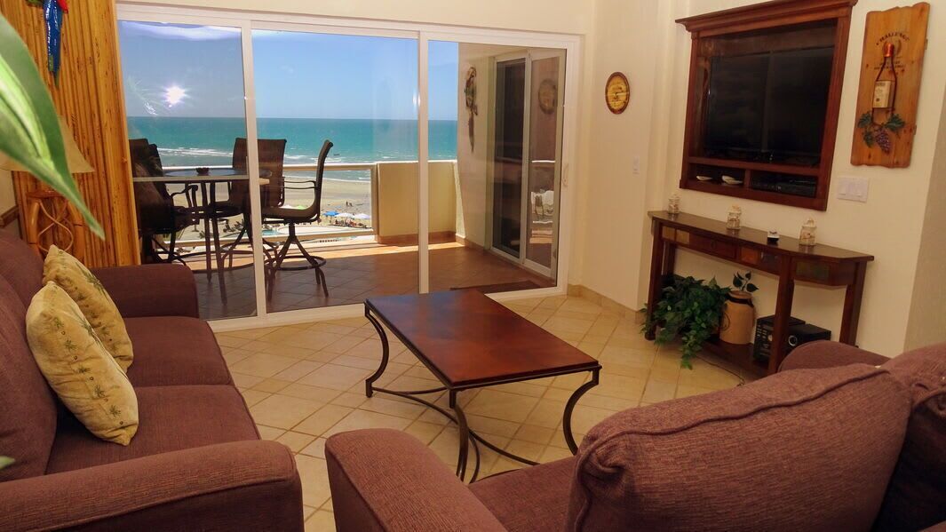 A living room with a view of the beach, a sliding glass door, a TV, and a small table.