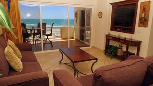 Uma sala de estar com vista para a praia, uma porta de vidro deslizante, uma televisão e uma pequena mesa.