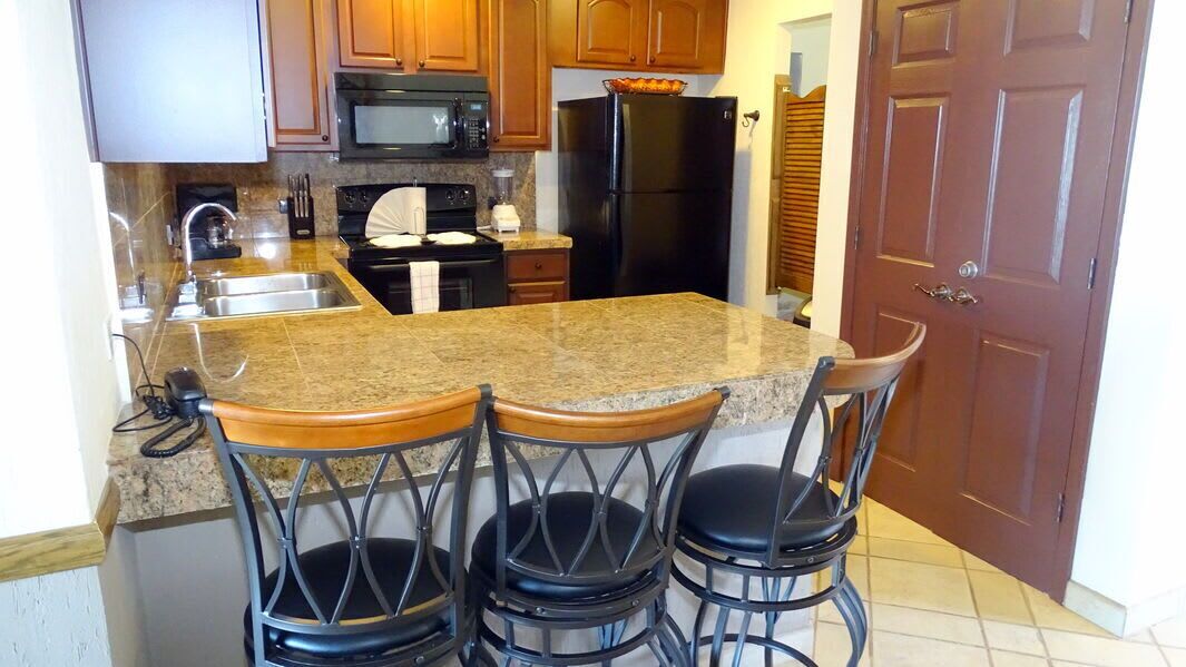 A kitchen with granite countertops, a black refrigerator, wooden cabinets, a microwave, and a sink. There are three bar stools with black seats.