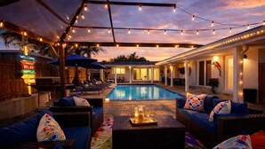 An outdoor pool area with lounge chairs and umbrellas, a covered seating area, and a sign with directional arrows. - Luxe Pool Retreat:  Heated Pool, Game Room, Sleeps 14 (Fort Lauderdale)