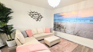 A modern living room with a sectional sofa, a pink rug, and a beach mural on the wall. - Fort Myers Coastal Retreat Hot Tub Yard Games (Fort Myers)