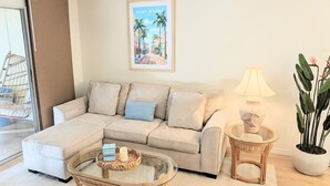 A living room with a beige sectional sofa, a glass coffee table, a wicker side table, a potted plant, and a framed picture of Fort Myers, Florida. - Retro Coastal Fort Myers Retreat Spa Fire Pit (Fort Myers)