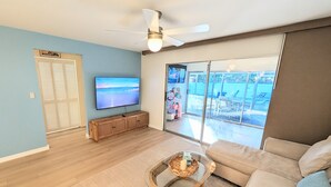 A modern living room with a flat-screen TV, a ceiling fan, a large sliding glass door to a balcony, and a wicker coffee table. - Retro Coastal Fort Myers Retreat Spa Fire Pit (Fort Myers)