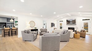 Een moderne woonkamer met grijze banken, een eethoek en een keuken op de achtergrond, plus een open haard.