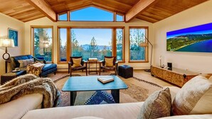 A spacious living room with a high ceiling, large windows offering a view of trees and mountains, a cozy sofa, a coffee table, and a flat-screen TV.