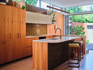 A modern kitchen with wooden cabinets, a large island with stools, and a view of the outdoors. - Northroy House / The Wemby (Fitzroy North)
