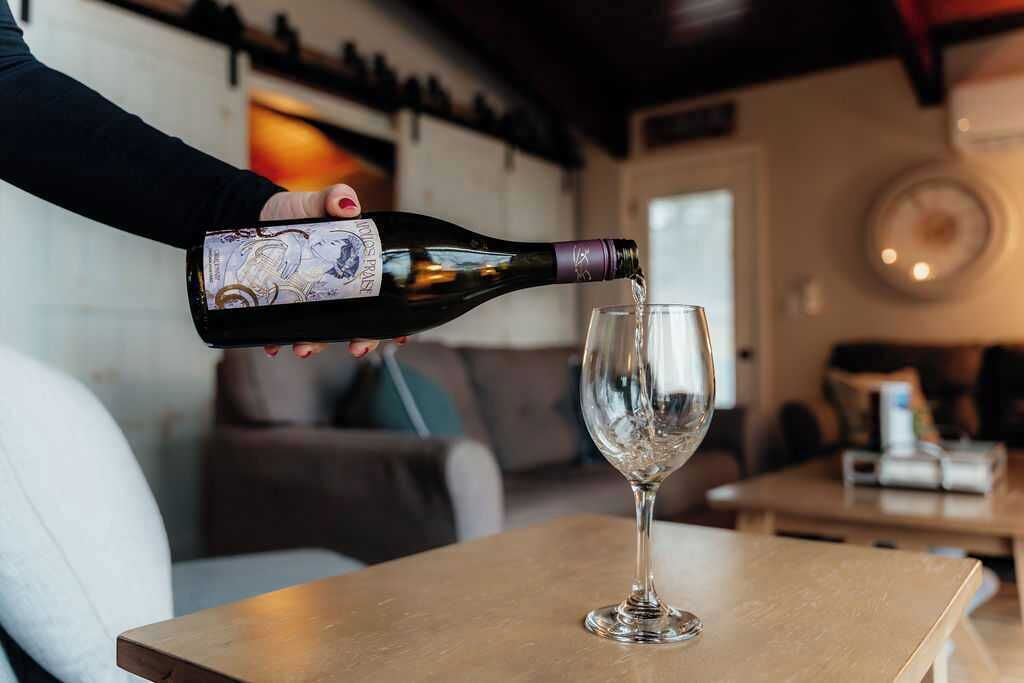 A person pouring wine from a bottle into a glass on a wooden table in a room with a couch and a wall clock.