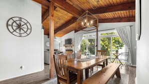 Un coin repas avec une table en bois, des chaises et un banc. On y trouve une décoration murale en forme de boussole, un lustre et une vue sur l’extérieur grâce à de grandes fenêtres.