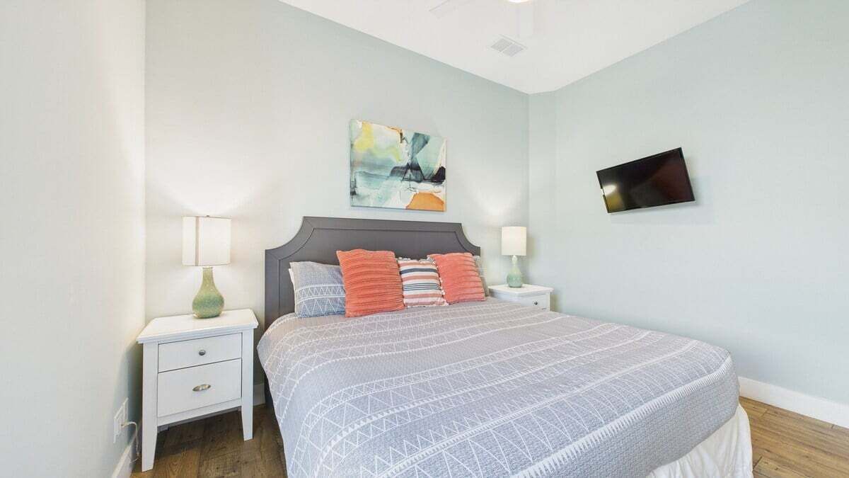 Un dormitorio con una cama, dos mesitas de noche, una mesilla auxiliar, un televisor montado en la pared y un cuadro.