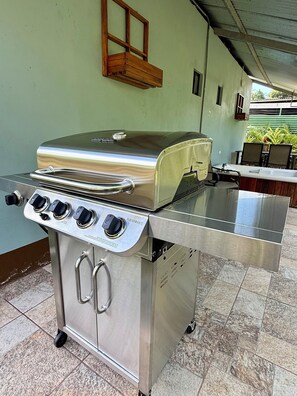 A stainless steel barbecue grill with four burners and a side cabinet, located in an outdoor dining area with tiled flooring and a green wall. - Cozy 2-bedroom house in delightful La Fortuna with AC (La fortuna)