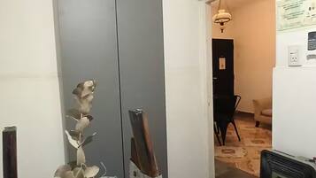 Una habitación con una puerta gris cerrada, un radiador, una mesita con un florero, y un pasillo que conduce a otra habitación con una silla negra y una lámpara.