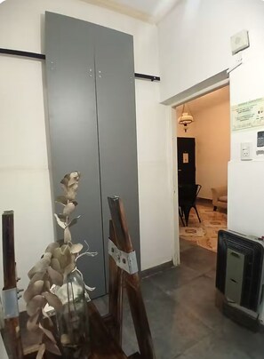A room with a closed grey door, a radiator, a small table with a vase, and a hallway leading to another room with a black chair and a lamp. - Serena Suipacha  (Mendoza)