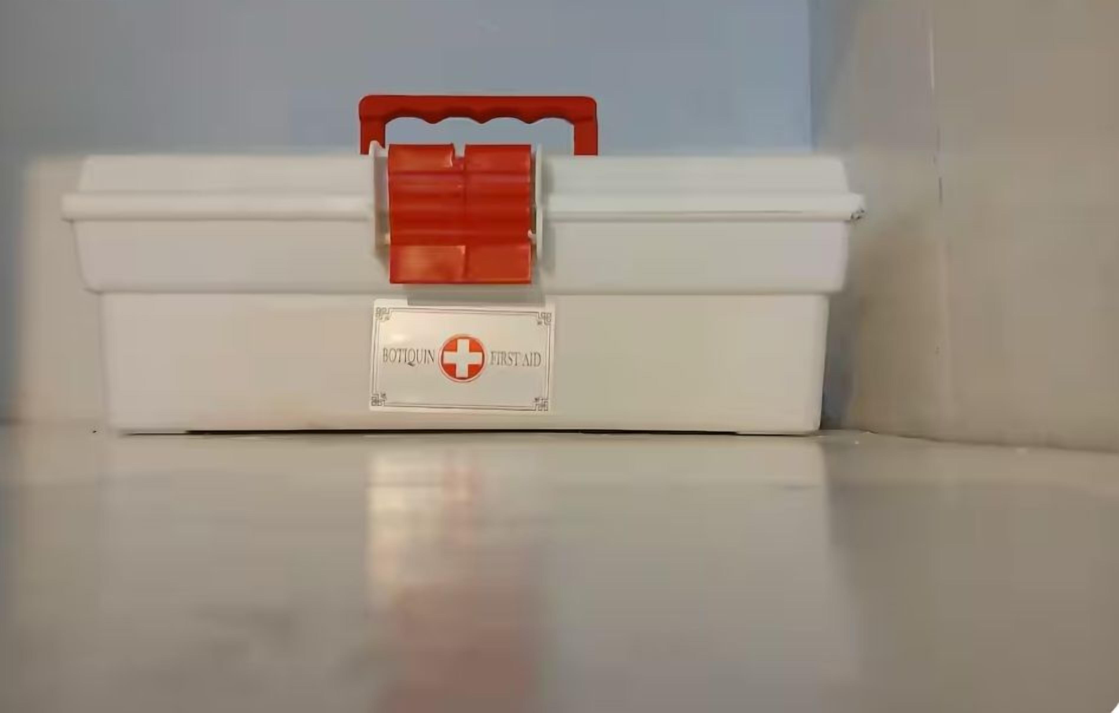 Un botiquín de primeros auxilios blanco con un asa roja y una etiqueta que dice 'BOTTQUIN FIRST AID'.