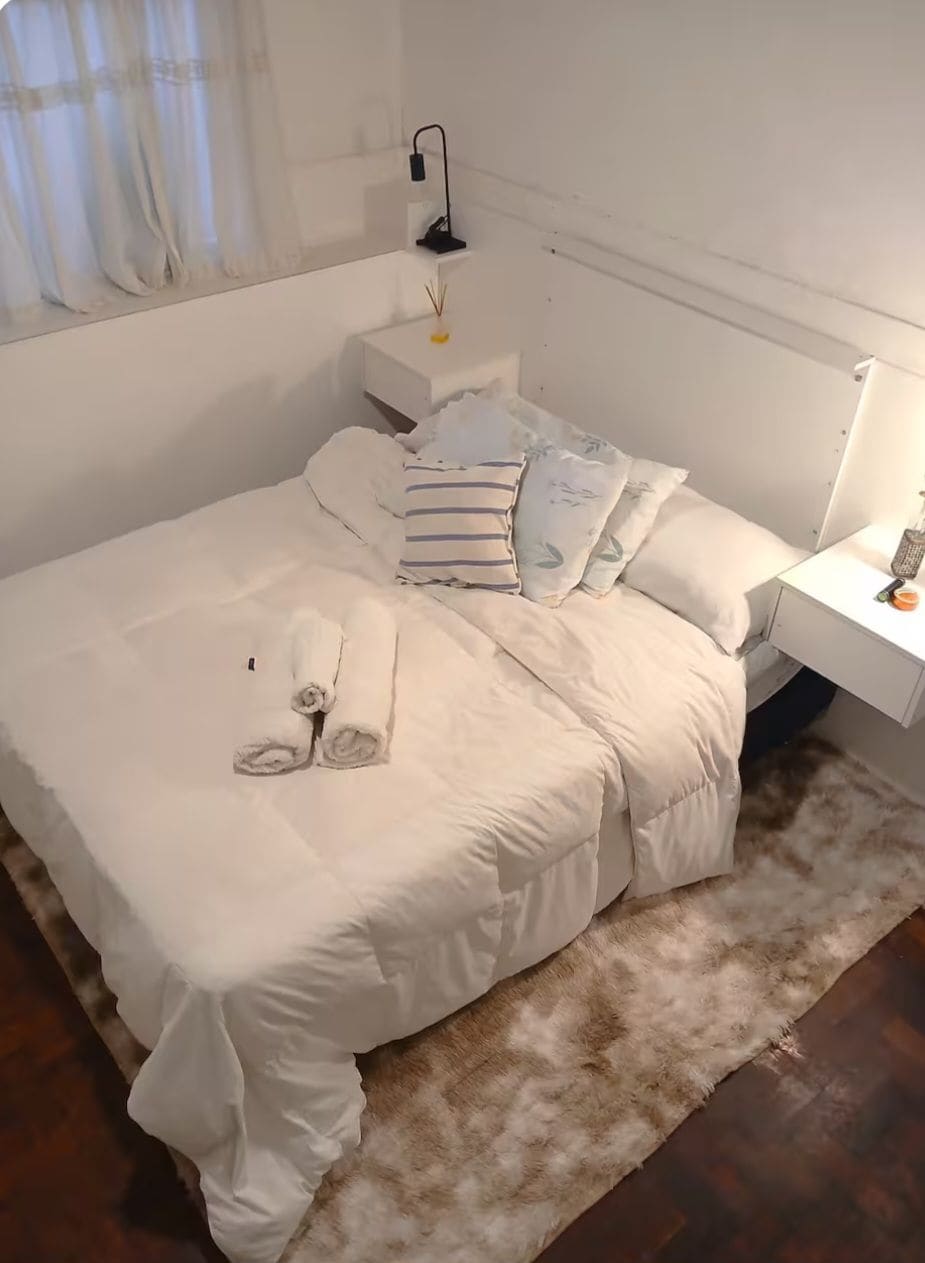 Un letto ben rifatto con un piumone bianco e cuscini, un comodino con una lampada e un tappeto morbido sul pavimento.