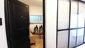 Una habitación con una puerta negra, una ventana con persianas, una pequeña mesa con sillas y suelo alfombrado.