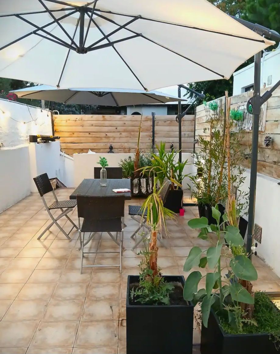Un patio al aire libre con mesa y sillas, rodeado de plantas en macetas y un paraguas grande.