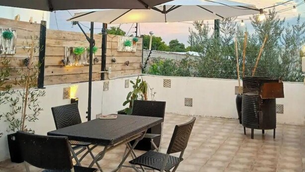 Un patio exterior con mesa y sillas bajo un paraguas grande, rodeado de plantas en macetas y una parrilla.