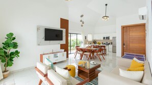 Un salon moderne avec un téléviseur à écran plat, un coin repas avec une table et des chaises en bois, une cuisine en arrière-plan et une grande porte en bois.