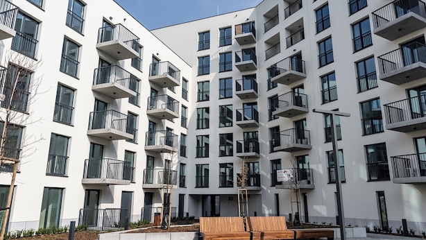 Un moderno edificio residenziale a più piani con balconi, un cortile centrale con una panchina in legno e cielo sereno.