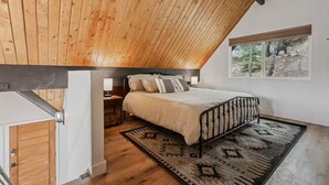 Quarto com teto inclinado, cama com colcha estampada, tapete e janela com vista para árvores.