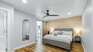 Un dormitorio moderno con suelo de madera, una cama con ropa de cama gris, un ventilador de techo y un espejo grande.