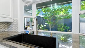 Una cocina moderna con un lavamanos grande, armarios blancos y vista a un árbol a través de ventanas amplias.