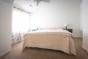 Ein Schlafzimmer mit Bett, Nachttisch, Lampe, Deckenventilator und einem Fenster mit Vorhängen.