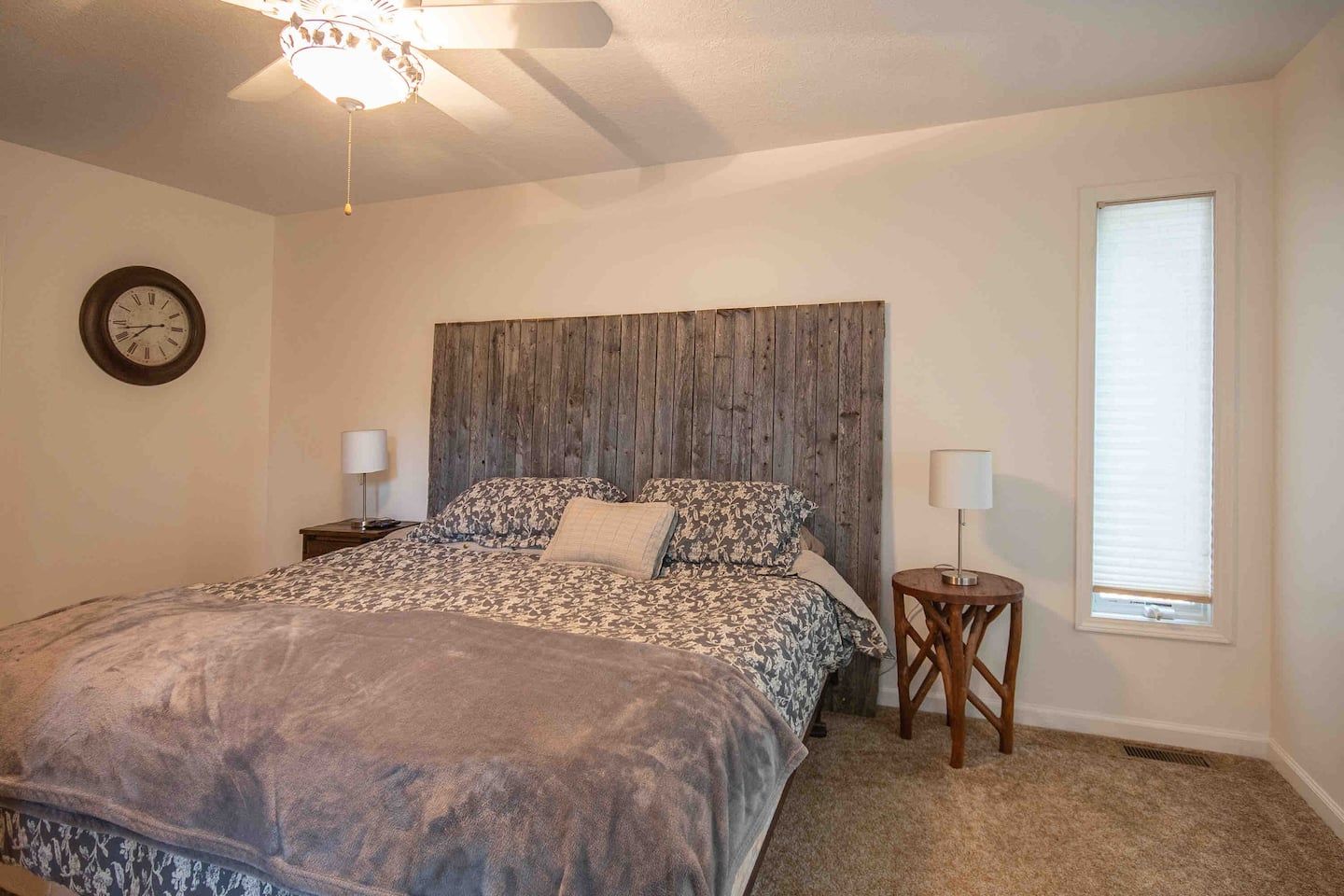A bedroom with a bed, a wooden headboard, a clock, two bedside tables with lamps, and a window with blinds.