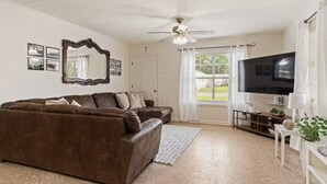 A living room with a brown leather couch, a ceiling fan, a large mirror, and a television. - The Rose 1 (Broussard)