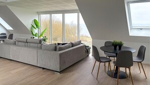Una sala de estar moderna con un sofá seccional gris, una mesa de comedor con sillas y ventanas amplias que ofrecen vistas a árboles y una masa de agua.