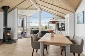 A modern interior with a wood-burning stove, a dining area with chairs, and a large window overlooking the sea. - Lovely holiday home panoramic view over the sea (Slagelse)