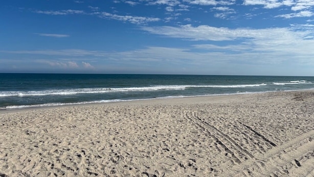 En sandstrand med fodaftryk, der fører ned mod vandet, en klar himmel og bløde bølger.