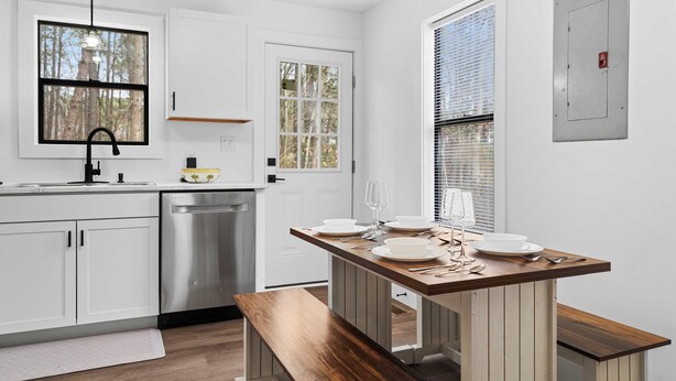 A kitchen with white cabinets, a stainless steel dishwasher, a wooden dining table with benches, and a window with a view of trees. - Minutes From UNC - 15 Min to Duke - Private Deck (Chapel Hill)